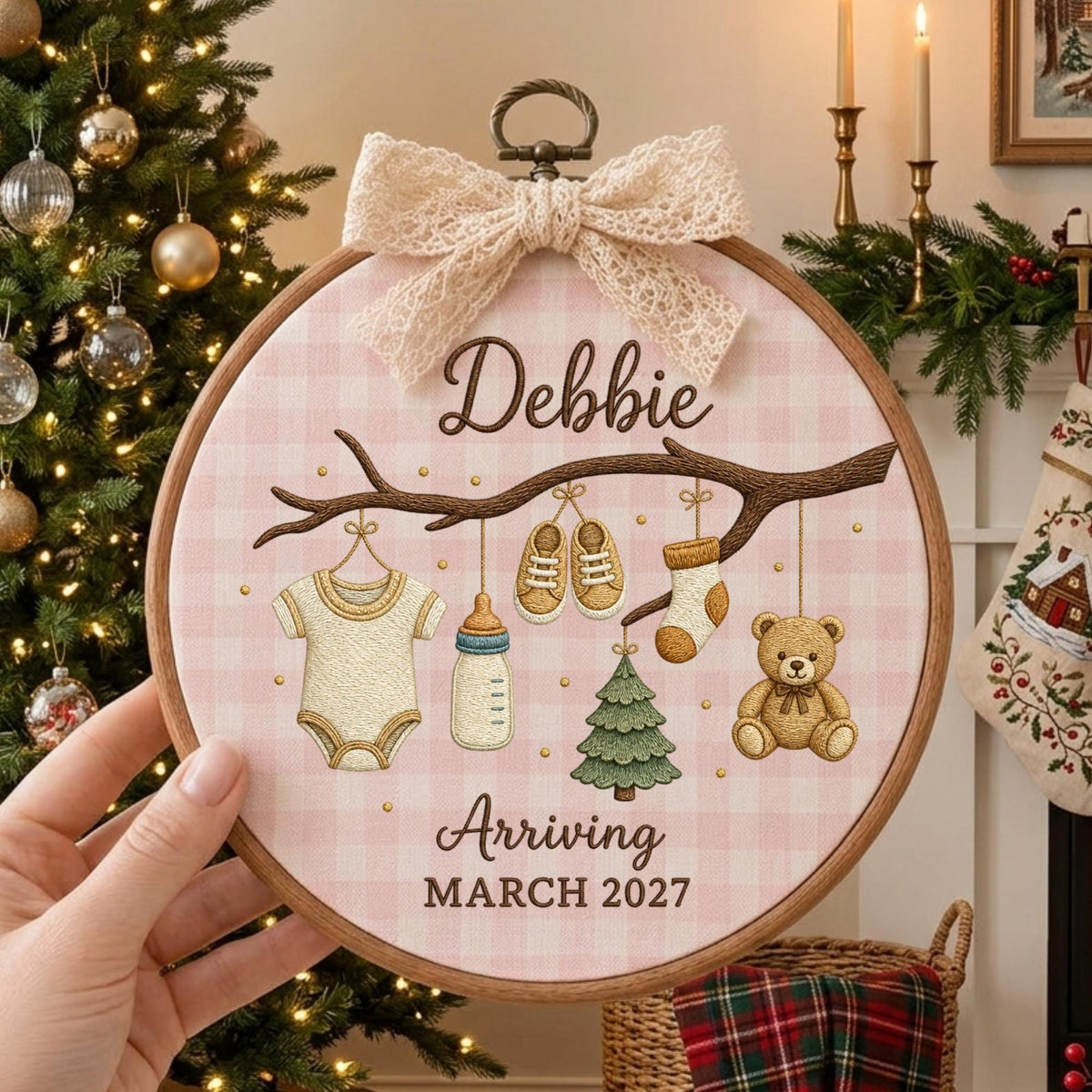 May include: A round wooden embroidery hoop featuring a pink gingham fabric background. The design includes a brown branch with a baby onesie, a bottle, shoes, a sock, a tree, and a teddy bear. Text reads: Debbie Arriving MARCH 2027.