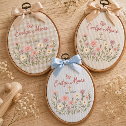 May include: Three oval wooden embroidery hoops featuring fabric with floral patterns, butterflies, and a cross. Each hoop displays the text Evelyn Moore and Child of God, topped with a satin ribbon bow in beige, light pink, or light blue.