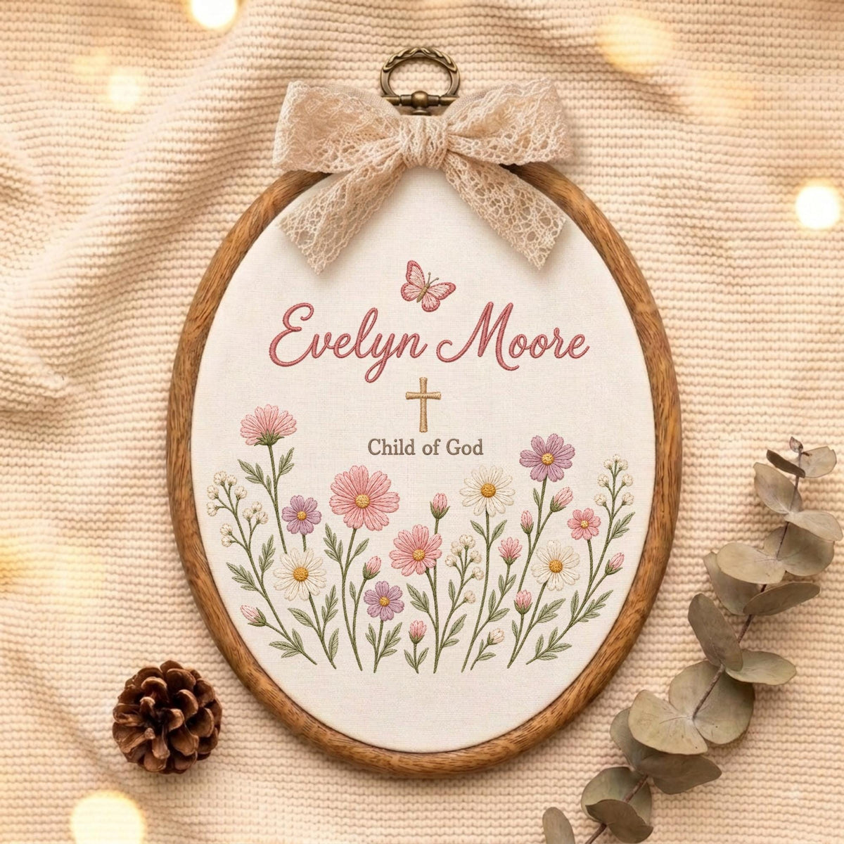 May include: An oval embroidery hoop featuring a cream fabric background with the embroidered name Evelyn Moore, a small butterfly, a cross, and the text Child of God, surrounded by pink and white flowers, topped with a beige lace bow.