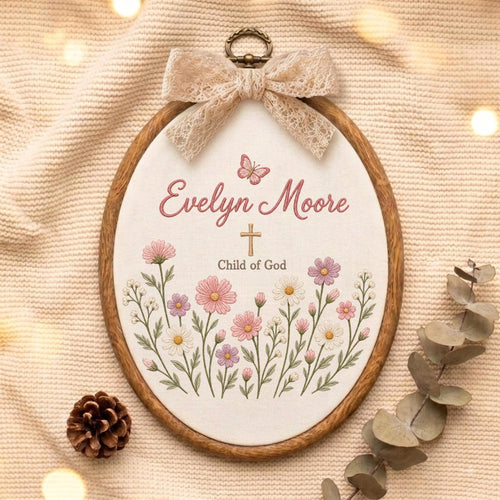 May include: An oval embroidery hoop featuring a cream fabric background with the embroidered name Evelyn Moore, a small butterfly, a cross, and the text Child of God, surrounded by pink and white flowers, topped with a beige lace bow.