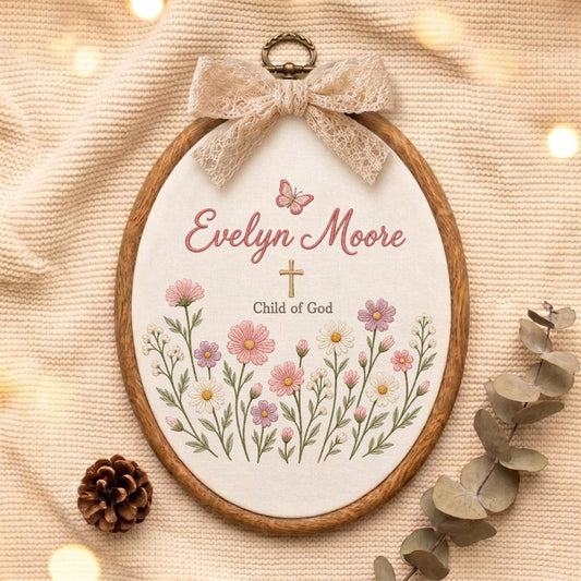May include: An oval embroidery hoop featuring a cream fabric background with the embroidered name Evelyn Moore, a small butterfly, a cross, and the text Child of God, surrounded by pink and white flowers, topped with a beige lace bow.