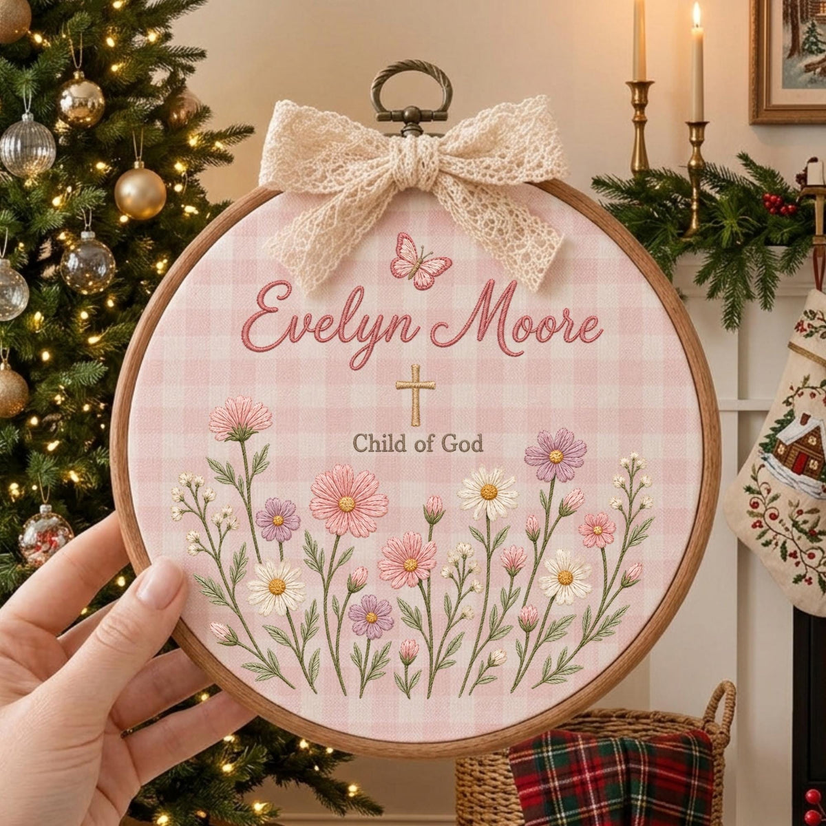 May include: A circular wooden embroidery hoop featuring a light pink gingham fabric background. The design includes embroidered pink and white flowers, a small butterfly, a gold cross, and the text Evelyn Moore Child of God, topped with a lace bow.