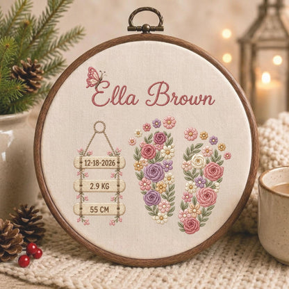 May include: A circular wooden embroidery hoop containing cream fabric with a hand-stitched design. The embroidery features the name Ella Brown, a small butterfly, two floral-filled baby footprints, and three hanging wooden signs reading 12-18-2026, 2.9 KG, and 55 CM.