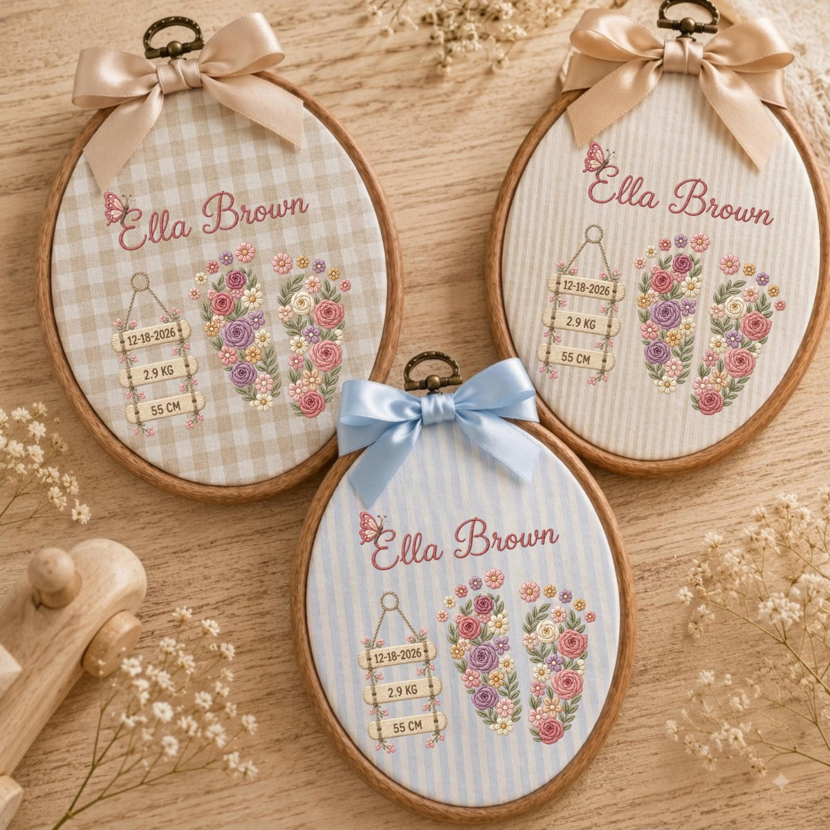May include: Three oval wooden embroidery hoops featuring fabric with the name Ella Brown, floral baby footprints, and birth details including 12-18-2026, 2.9 kg, and 55 cm. Each hoop is topped with a satin ribbon bow in beige or light blue.