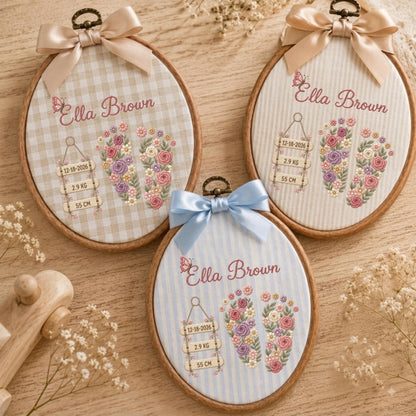 May include: Three oval wooden embroidery hoops featuring fabric with the name Ella Brown, floral baby footprints, and birth details including 12-18-2026, 2.9 kg, and 55 cm. Each hoop is topped with a satin ribbon bow in beige or light blue.