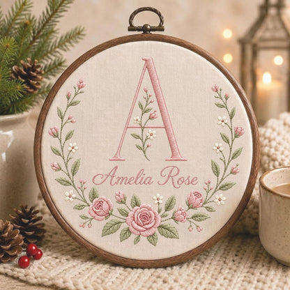 May include: A circular wooden embroidery hoop containing cream-colored fabric with a hand-stitched design. The embroidery features a large pink letter A, the name Amelia Rose in cursive script, and a floral wreath of pink roses and green leaves.