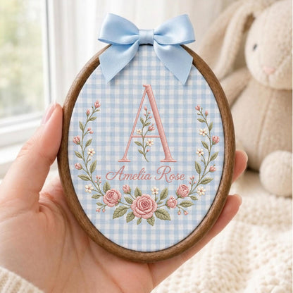 May include: An oval wooden embroidery hoop containing light blue and white gingham fabric. The fabric features an embroidered pink letter A, the name Amelia Rose in pink script, and a floral wreath of pink roses and green leaves with a light blue bow.