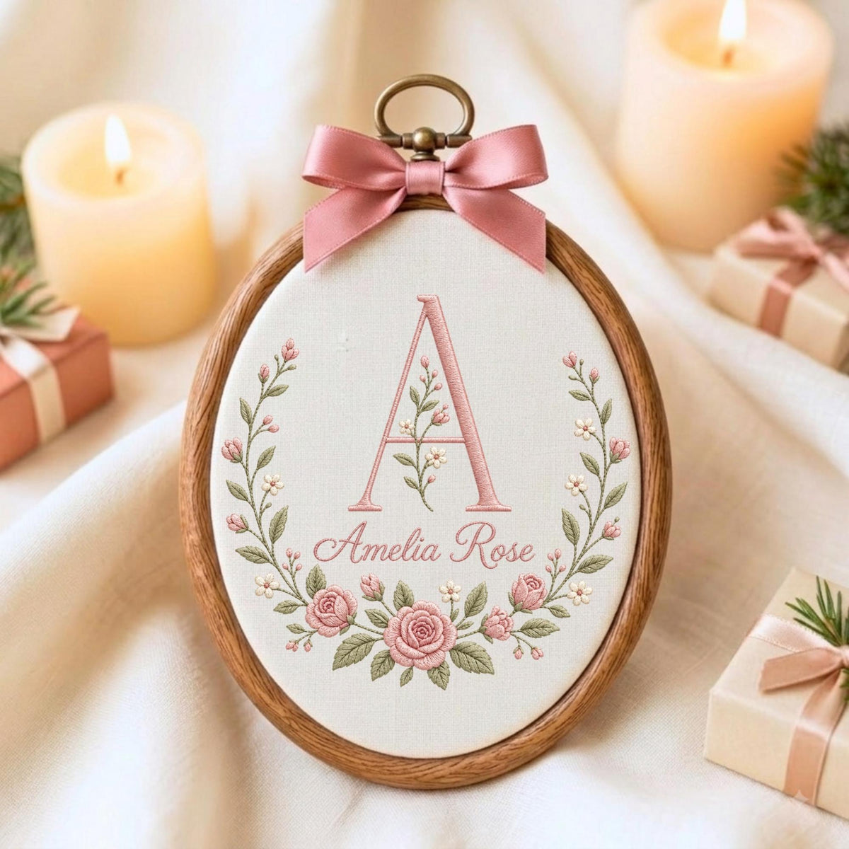 May include: An oval wooden embroidery hoop containing white fabric with a pink embroidered letter A and the name Amelia Rose. The design features a floral wreath of pink roses and green leaves. A pink satin ribbon bow is attached to the top.