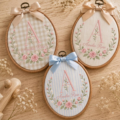 May include: Three oval wooden embroidery hoops featuring fabric backgrounds with embroidered floral wreaths, the letter A, and the name Amelia Rose. Two hoops have beige fabric with gingham or striped patterns and beige bows, one has blue stripes and a blue bow.
