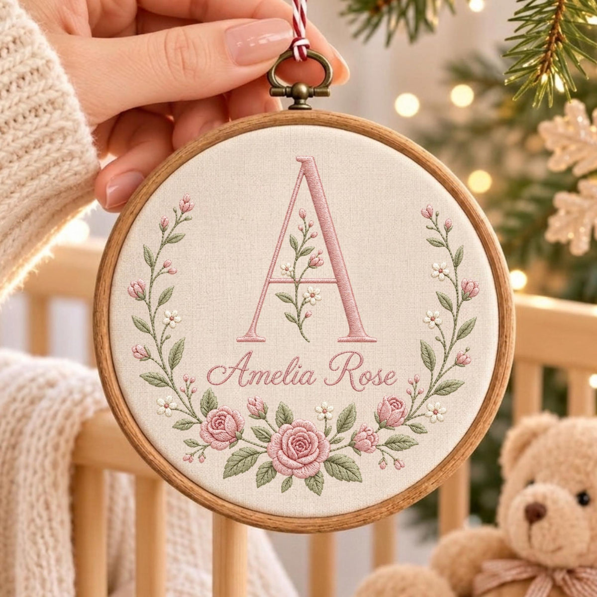 May include: A circular wooden embroidery hoop containing off-white fabric with a hand-stitched design. The design features a large pink letter A, the name Amelia Rose in cursive script, and a floral wreath of pink roses and green leaves.
