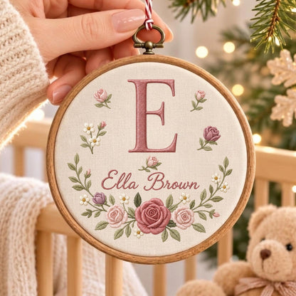 May include: A circular wooden embroidery hoop containing off-white fabric with a dusty rose embroidered letter E, the name Ella Brown in cursive script, and a decorative wreath of pink roses, rosebuds, and small white flowers with green leaves.