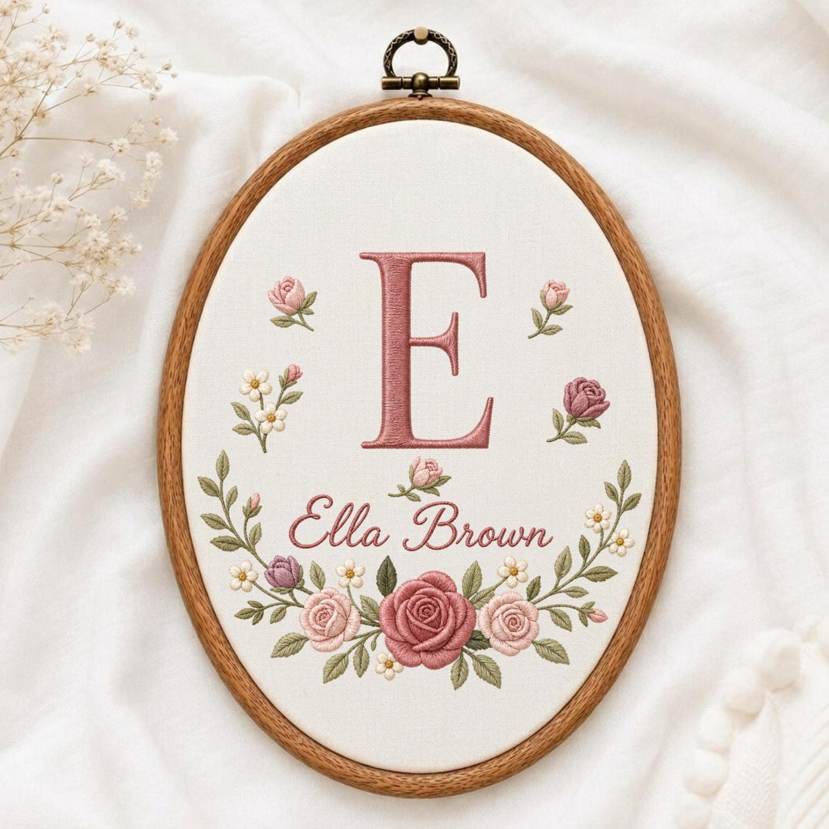 May include: An oval wooden embroidery hoop containing white fabric with a hand-stitched design. The embroidery features a large pink letter E, the name Ella Brown in cursive script, and a floral wreath of pink roses and small white flowers.