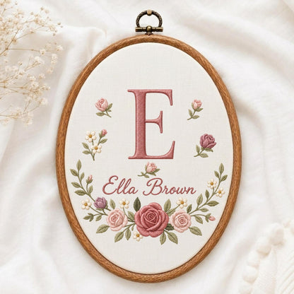 May include: An oval wooden embroidery hoop containing white fabric with a hand-stitched design. The embroidery features a large pink letter E, the name Ella Brown in cursive script, and a floral wreath of pink roses and small white flowers.