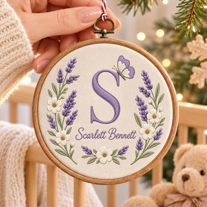 May include: A circular wooden embroidery hoop containing off-white fabric with a hand-stitched design. The design features a large purple letter S, a small butterfly, and floral sprigs of lavender and white flowers with the name Scarlett Bennett.
