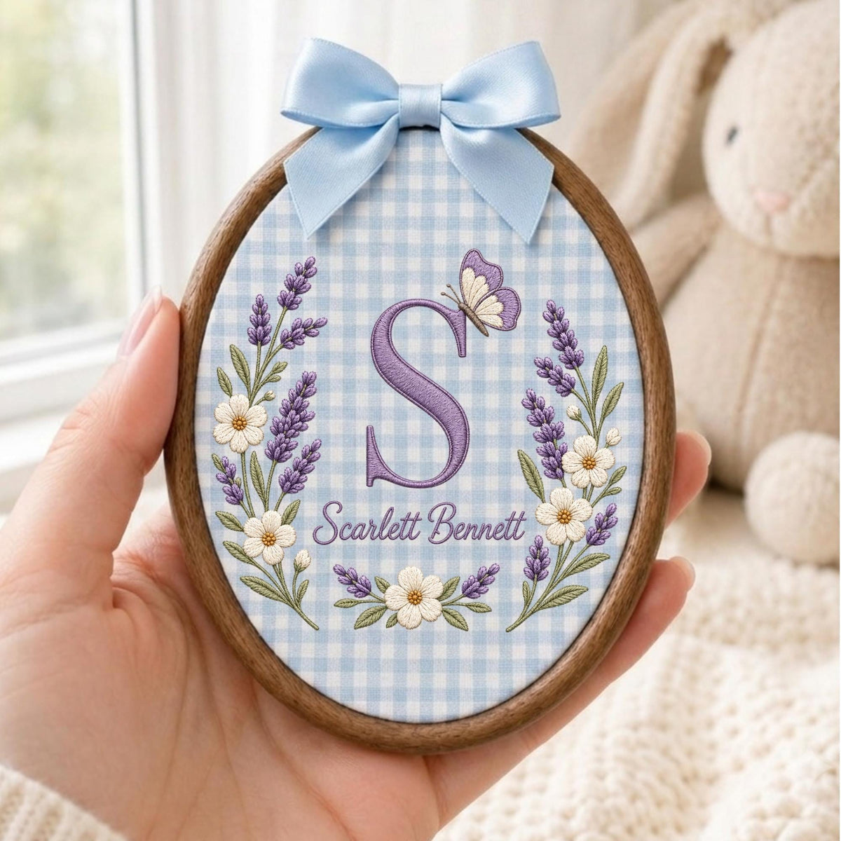 May include: An oval wooden embroidery hoop containing fabric with a light blue and white gingham pattern. The fabric features an embroidered purple letter S, a butterfly, lavender sprigs, white flowers, and the text Scarlett Bennett, topped with a light blue satin bow.