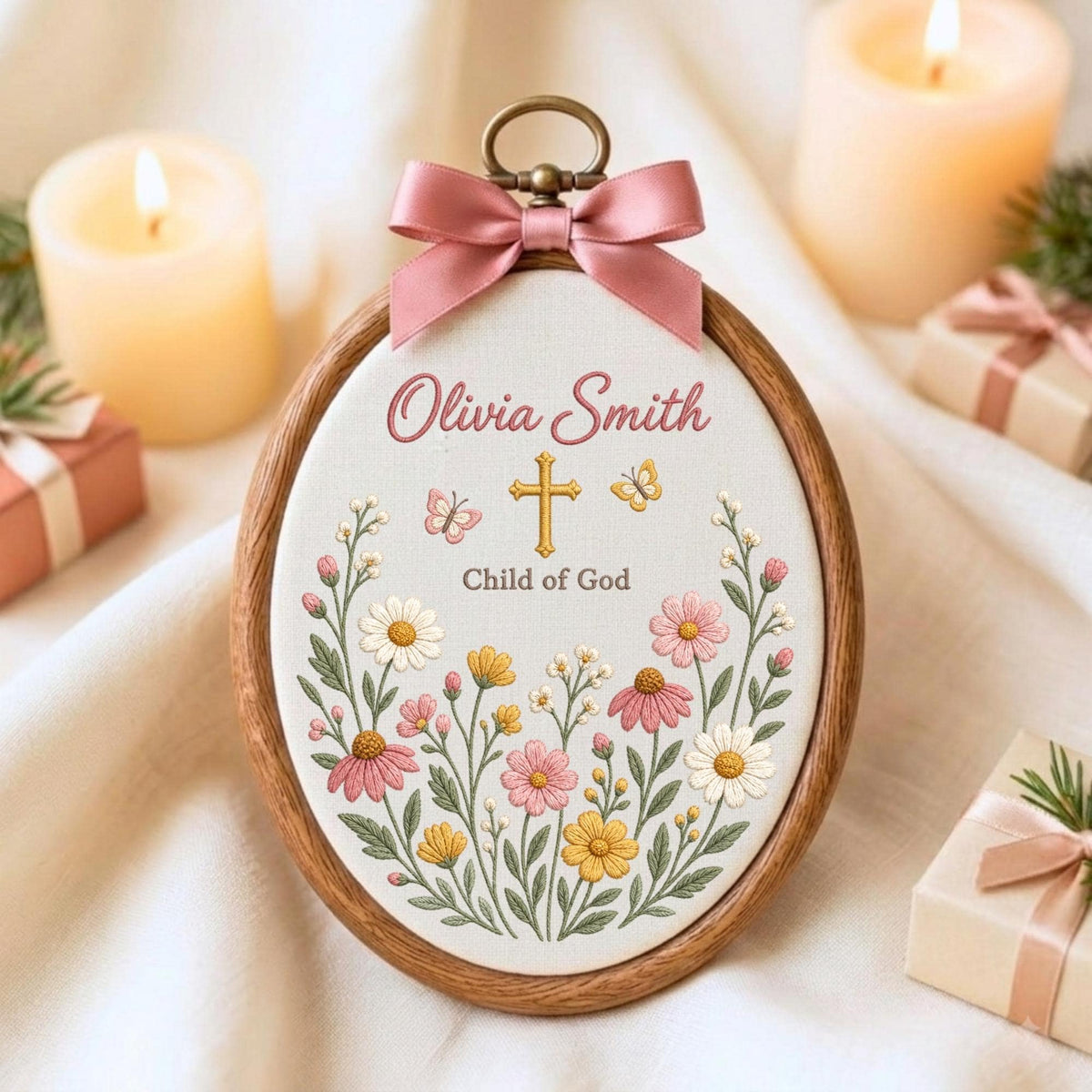 May include: An oval embroidery hoop featuring a white fabric center with the embroidered text Olivia Smith and Child of God, a gold cross, two butterflies, and colorful wildflowers in pink, yellow, and white, topped with a pink satin bow.