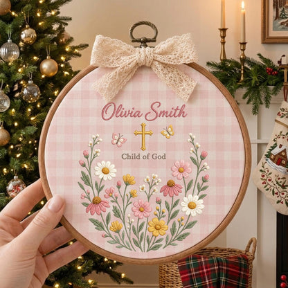 May include: A circular wooden embroidery hoop featuring a light pink gingham fabric background. The design includes the name Olivia Smith, a gold cross, the text Child of God, and a floral wreath with butterflies, topped with a lace bow.