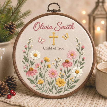May include: A circular wooden embroidery hoop containing fabric with the embroidered name Olivia Smith, a gold cross, and the text Child of God. The design features colorful flowers in shades of pink, yellow, and white, with two small butterflies.