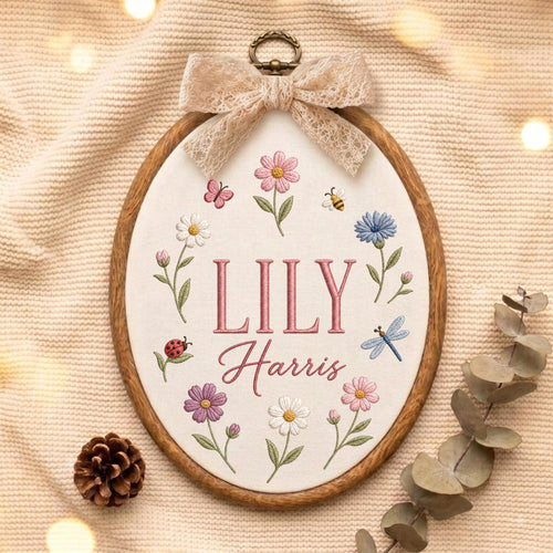 May include: An oval embroidery hoop featuring the name LILY Harris stitched in pink thread. The fabric is decorated with embroidered flowers in pink, white, blue, and purple, along with a ladybug, a butterfly, a bee, and a dragonfly. A beige lace bow is attached to the top.
