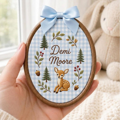 May include: An oval wooden embroidery hoop featuring a light blue and white gingham fabric background. The fabric is embroidered with the name Demi Moore, a brown fawn, green pine trees, acorns, and leafy branches. A light blue satin bow is attached.