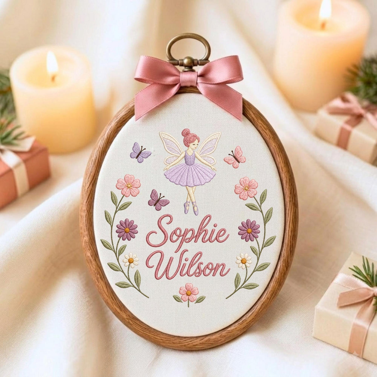 May include: An oval wooden embroidery hoop containing white fabric with a pink satin bow at the top. The embroidery features a fairy in a purple dress, butterflies, flowers, and the name Sophie Wilson in pink cursive script lettering.