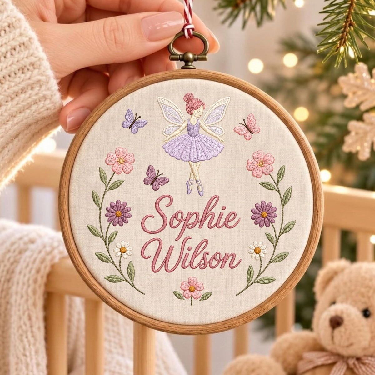 May include: A round wooden embroidery hoop containing off-white fabric with a fairy in a purple dress, butterflies, and flowers. The text "Sophie Wilson" is embroidered in pink script in the center. A red and white string hangs from the top.