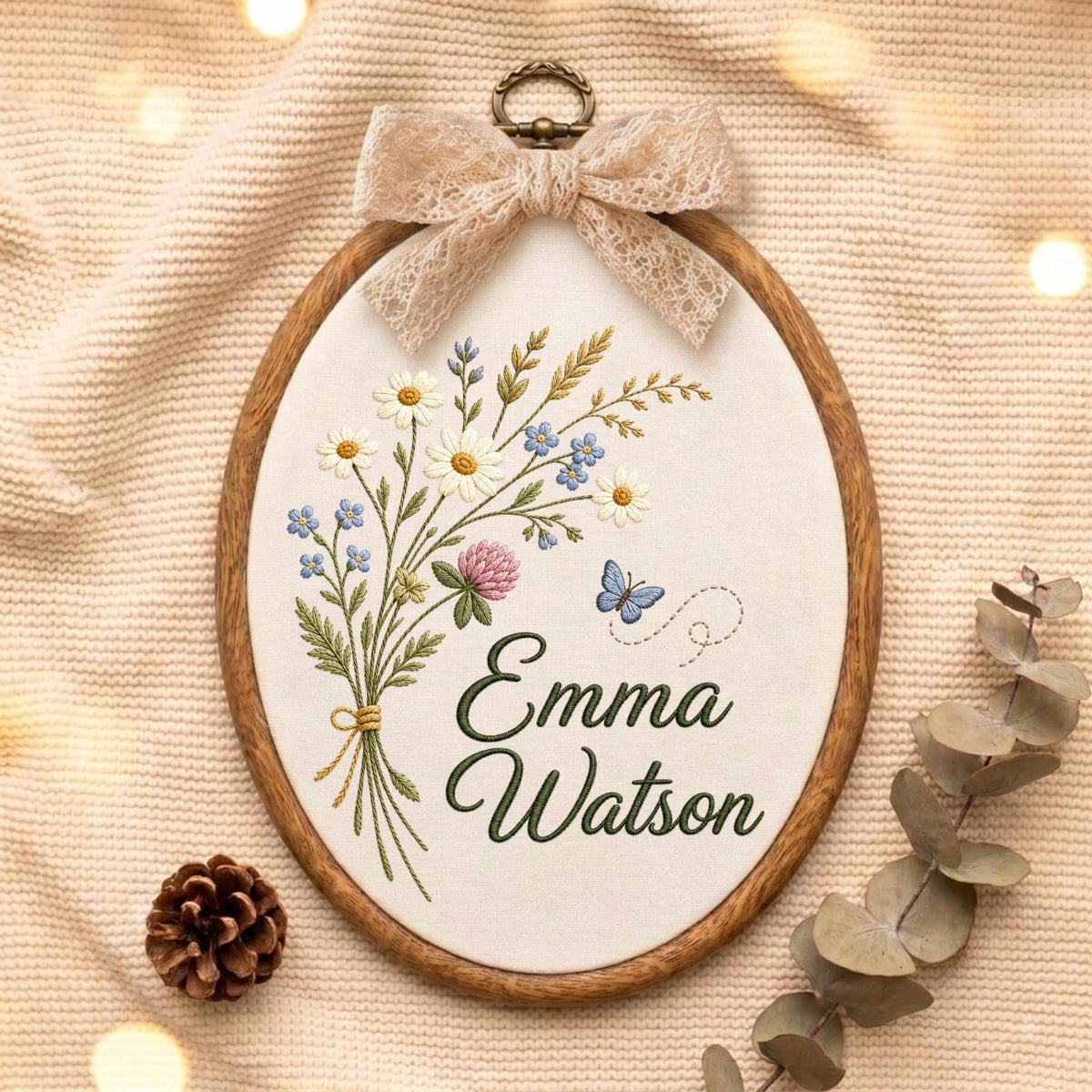 May include: An oval wooden embroidery hoop containing white fabric with a hand-stitched bouquet of wildflowers, a small blue butterfly, and the name Emma Watson in dark green cursive script, topped with a decorative beige lace bow.