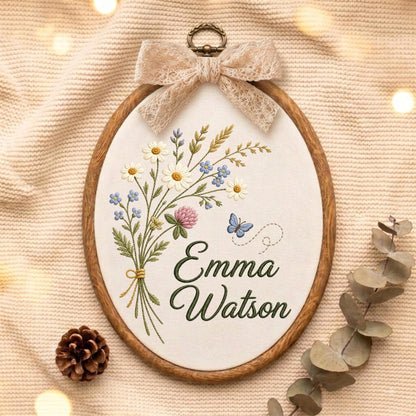 May include: An oval wooden embroidery hoop containing white fabric with a hand-stitched bouquet of wildflowers, a small blue butterfly, and the name Emma Watson in dark green cursive script, topped with a decorative beige lace bow.