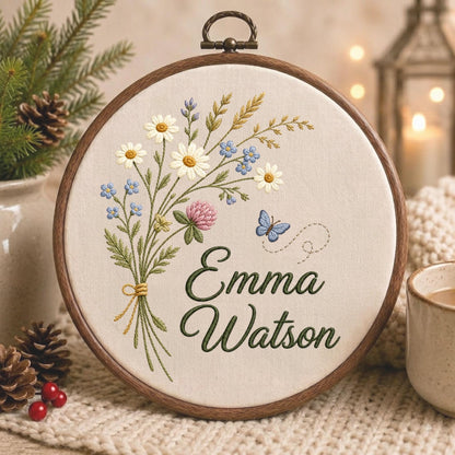 May include: A circular wooden embroidery hoop containing off-white fabric with a hand-stitched bouquet of daisies, clover, and blue wildflowers. A small blue butterfly is embroidered to the right, and the name Emma Watson is stitched in dark green.