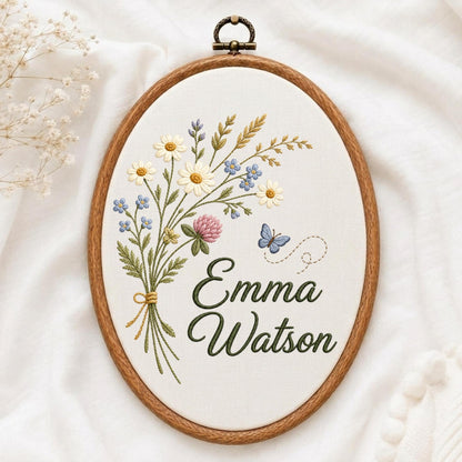 May include: An oval wooden embroidery hoop containing white fabric with a hand-stitched bouquet of wildflowers, including daisies, clover, and blue blossoms. A small blue butterfly is stitched nearby. The name Emma Watson is embroidered in green.
