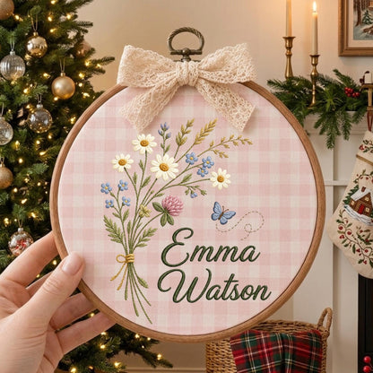 May include: A round wooden embroidery hoop featuring a pink and white gingham fabric background. The design includes an embroidered bouquet of daisies and wildflowers with a small blue butterfly, and the name Emma Watson in dark green script.