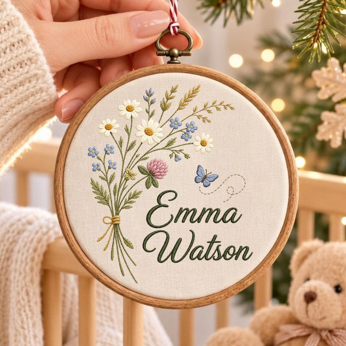 May include: A circular wooden embroidery hoop containing off-white fabric with a stitched bouquet of daisies, clover, and greenery. A small blue butterfly is embroidered to the right. The name Emma Watson is stitched in dark green cursive script.