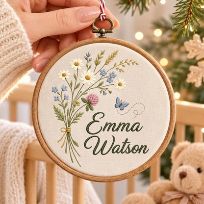 May include: A circular wooden embroidery hoop containing off-white fabric with a stitched bouquet of daisies, clover, and greenery. A small blue butterfly is embroidered to the right. The name Emma Watson is stitched in dark green cursive script.
