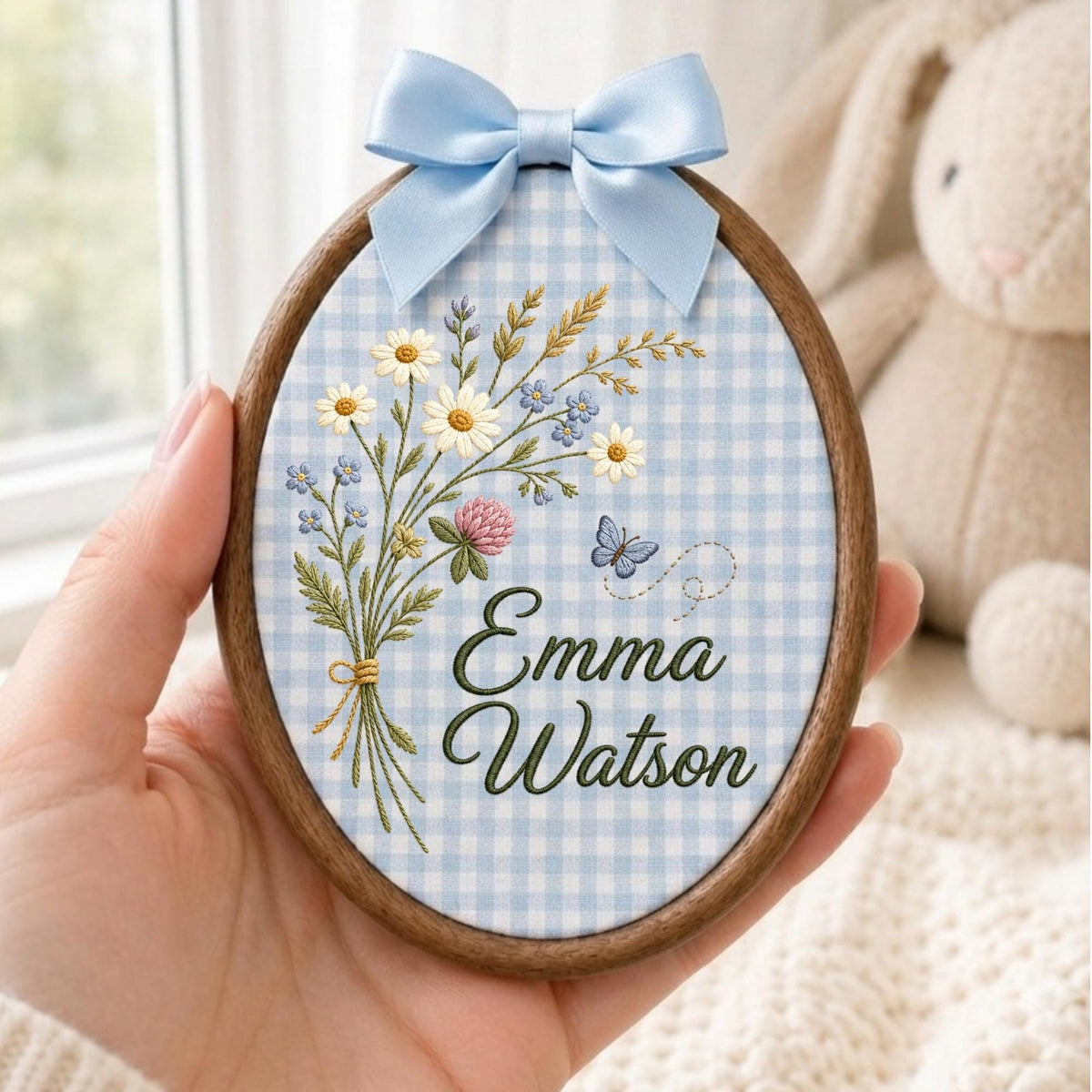 May include: An oval wooden embroidery hoop containing light blue and white gingham fabric. The fabric features an embroidered bouquet of wildflowers, a small blue butterfly, and the dark green text Emma Watson. A light blue satin bow is attached.