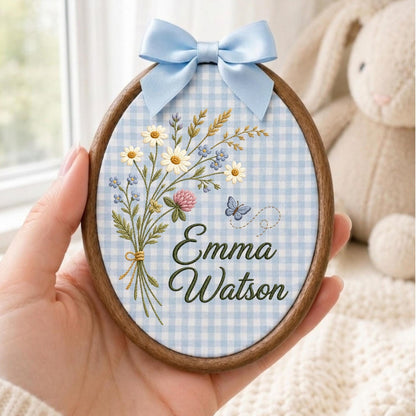 May include: An oval wooden embroidery hoop containing light blue and white gingham fabric. The fabric features an embroidered bouquet of wildflowers, a small blue butterfly, and the dark green text Emma Watson. A light blue satin bow is attached.