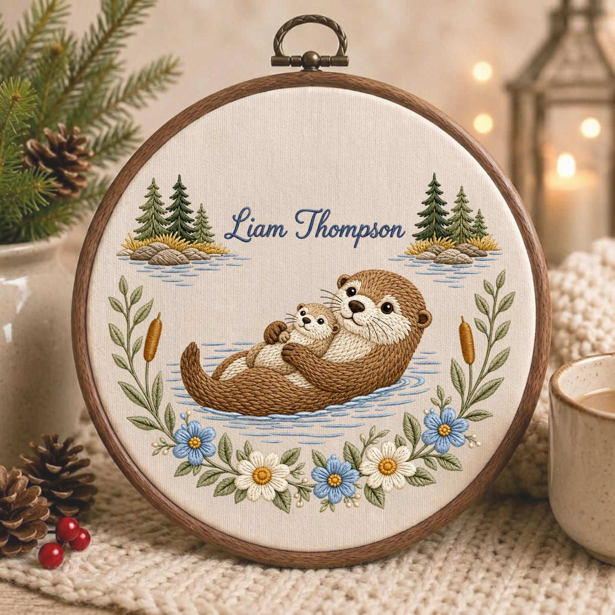 May include: A circular embroidery hoop featuring a stitched design of a brown otter holding a pup in blue water. The scene includes green pine trees, cattails, and a floral border of blue and white flowers. The text Liam Thompson is embroidered.