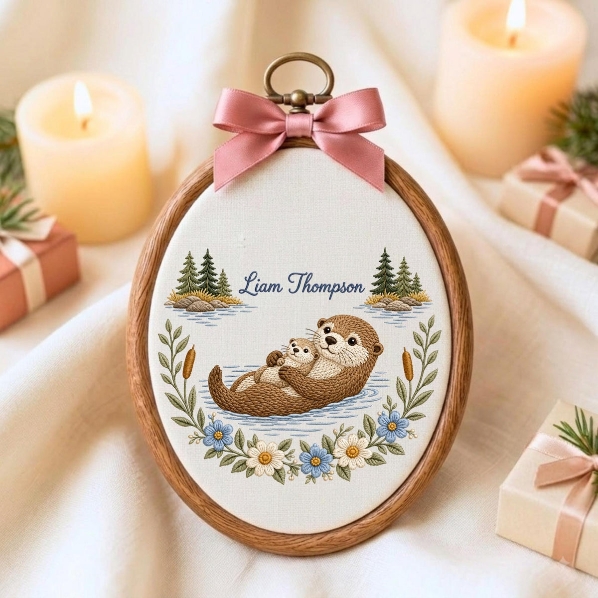 May include: An oval wooden embroidery hoop containing fabric with a stitched illustration of two brown otters floating in water. The text Liam Thompson is embroidered above the otters, surrounded by pine trees, cattails, and flowers, with a pink bow.