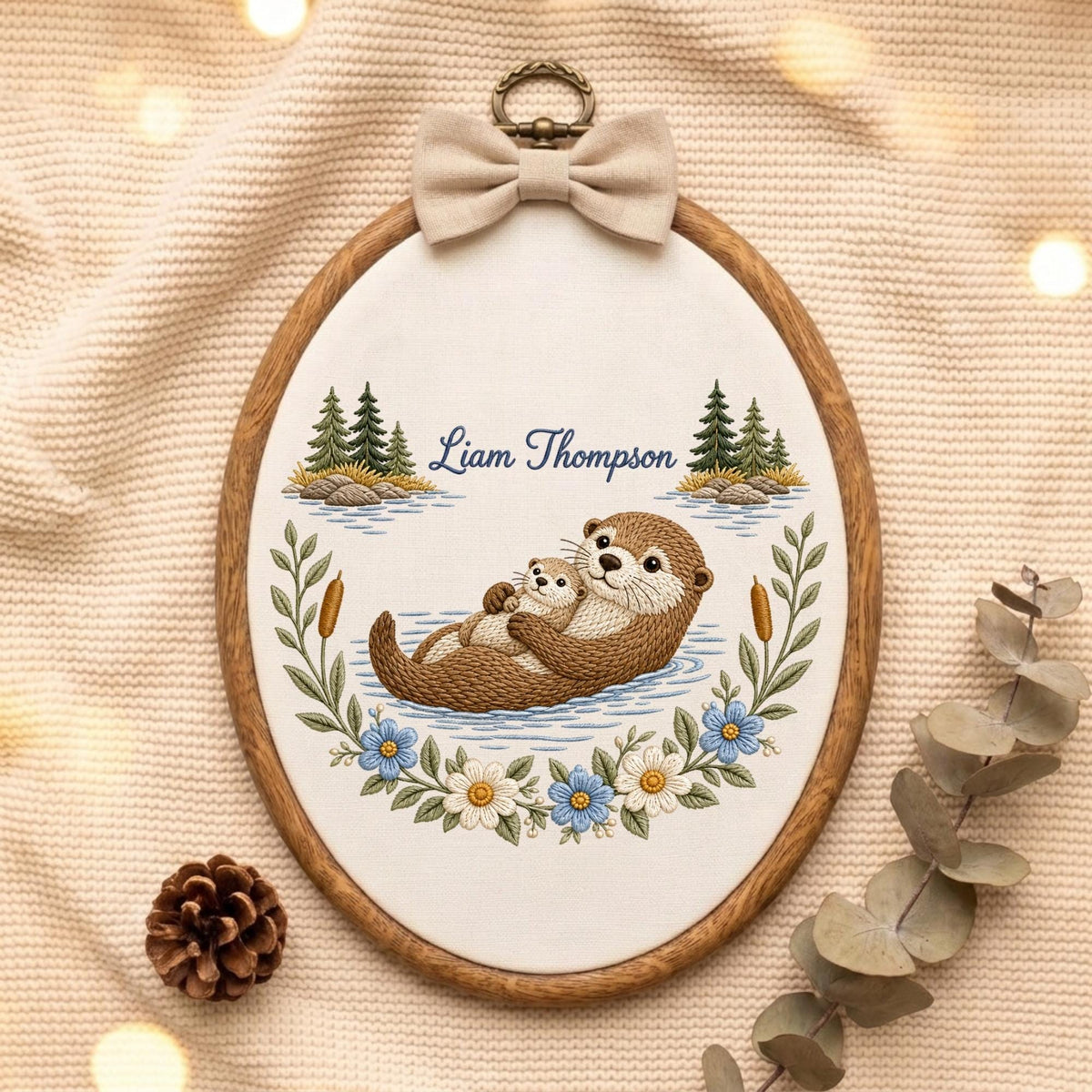 May include: An oval wooden embroidery hoop containing a fabric panel with an embroidered scene of two brown otters floating in water. The text Liam Thompson is stitched above the otters, flanked by pine trees, cattails, and blue and white flowers.