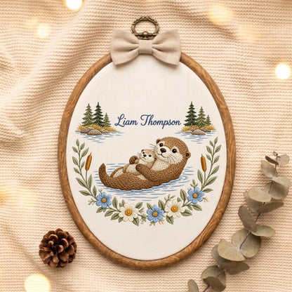 May include: An oval wooden embroidery hoop containing a fabric panel with an embroidered scene of two brown otters floating in water. The text Liam Thompson is stitched above the otters, flanked by pine trees, cattails, and blue and white flowers.