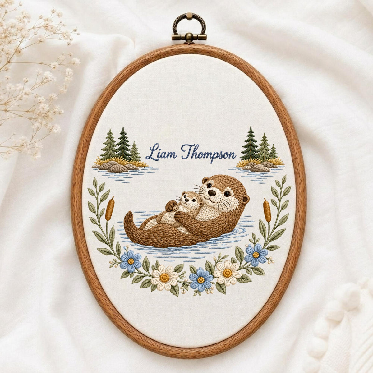 May include: An oval embroidery hoop featuring a brown otter holding a smaller otter on a light fabric background. The design includes embroidered pine trees, cattails, and blue and white flowers. The text Liam Thompson is stitched in blue.