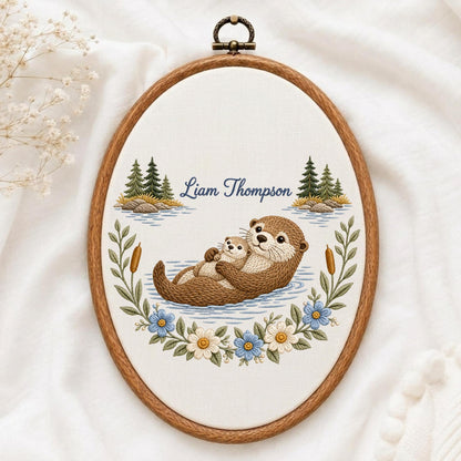 May include: An oval embroidery hoop featuring a brown otter holding a smaller otter on a light fabric background. The design includes embroidered pine trees, cattails, and blue and white flowers. The text Liam Thompson is stitched in blue.