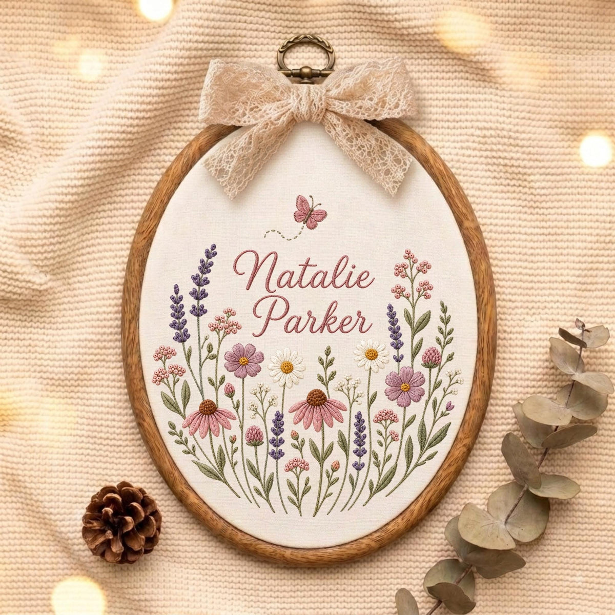 May include: An oval wooden embroidery hoop containing white fabric with a floral design featuring lavender, daisies, and pink wildflowers. The text Natalie Parker is embroidered in the center with a small pink butterfly above. A beige lace bow is attached.
