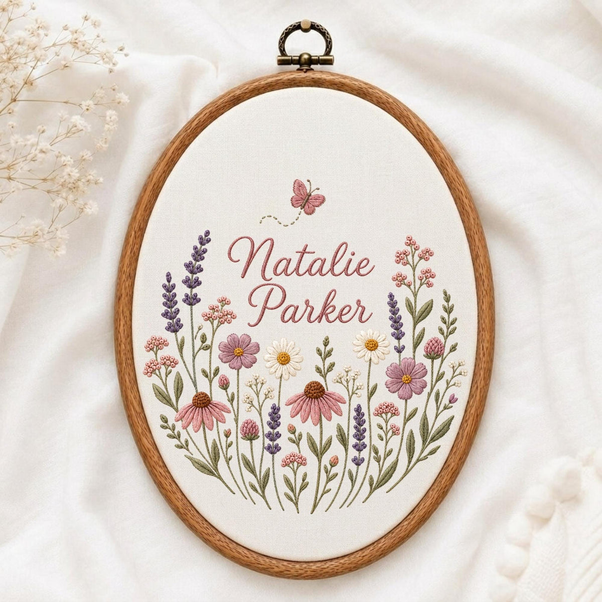 May include: An oval embroidery hoop containing white fabric with a floral design featuring purple, pink, and white wildflowers. The text "Natalie Parker" is embroidered in the center in a script font, with a small pink butterfly above the name.