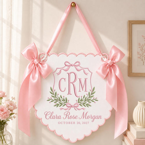 May include: A white scalloped wall hanging featuring pink embroidered text that reads Clara Rose Morgan and October 20, 2027, with a central monogram CRM, green leaf accents, and two large pink satin bows attached to the top corners with a ribbon.