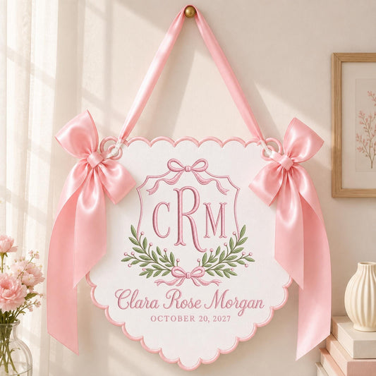 May include: A white scalloped wall hanging featuring pink embroidered text that reads Clara Rose Morgan and October 20, 2027, with a central monogram CRM, green leaf accents, and two large pink satin bows attached to the top corners with a ribbon.