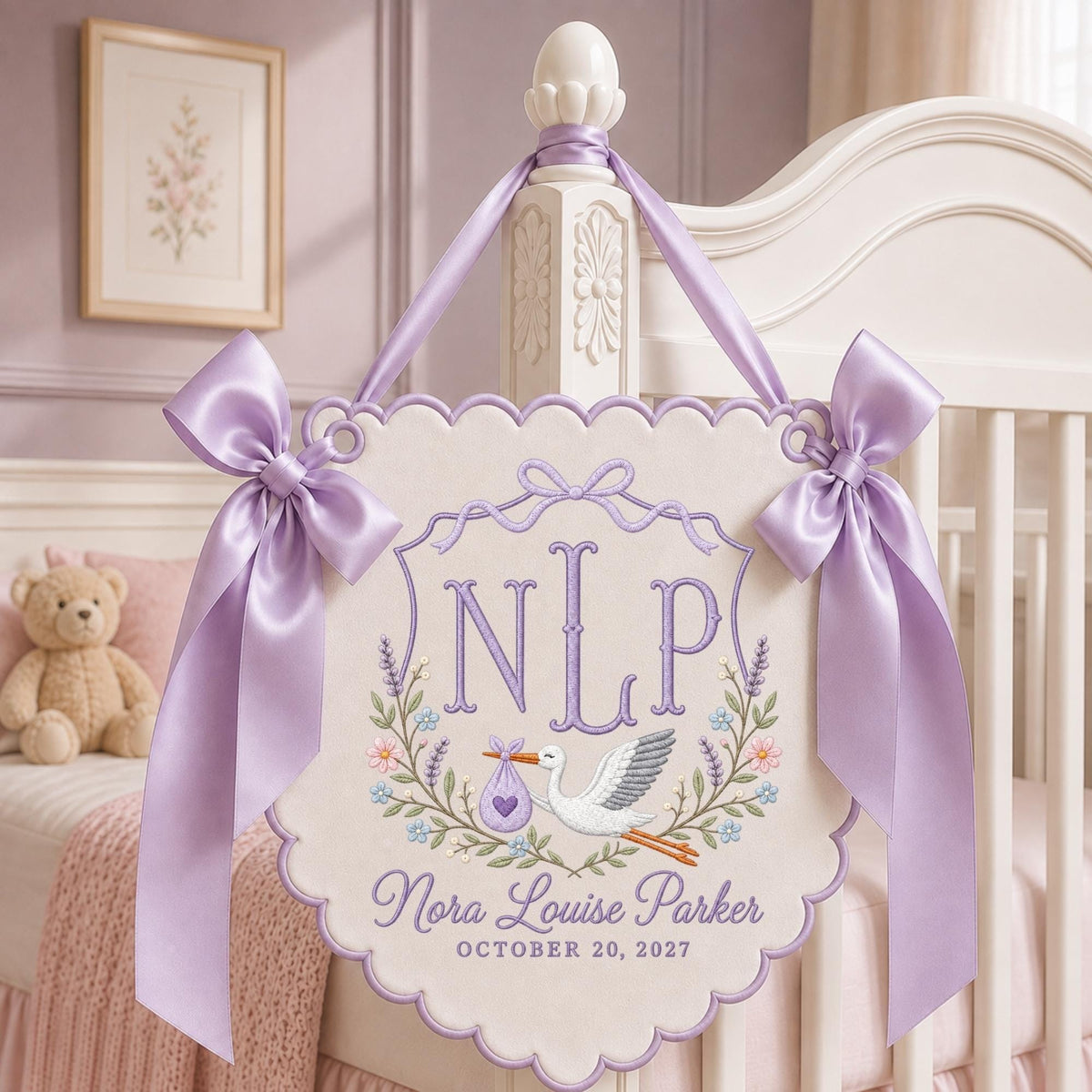 May include: A cream-colored scalloped wall hanging featuring lavender embroidery of the initials NLP, a stork carrying a purple bundle, and floral accents. The text reads Nora Louise Parker and OCTOBER 20, 2027, accented by two large lavender satin bows.
