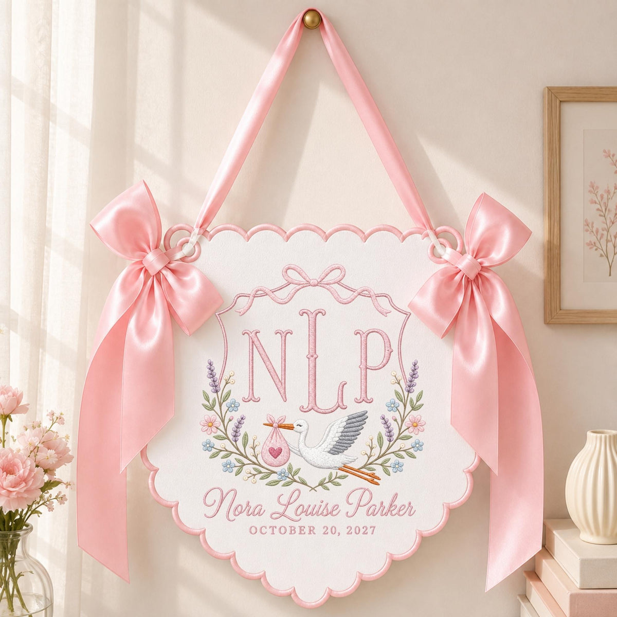 May include: A white scalloped wall hanging featuring pink satin ribbon bows and a pink embroidered design. The embroidery includes the monogram NLP, a stork carrying a pink bundle, floral accents, and the text Nora Louise Parker October 20, 2027.