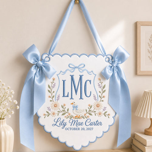 May include: A white scalloped wall hanging banner featuring blue embroidery of the monogram LMC, a small duck wearing a blue bow, and floral accents. The text reads Lily Mae Carter and OCTOBER 20, 2027. It is suspended by a light blue satin ribbon.