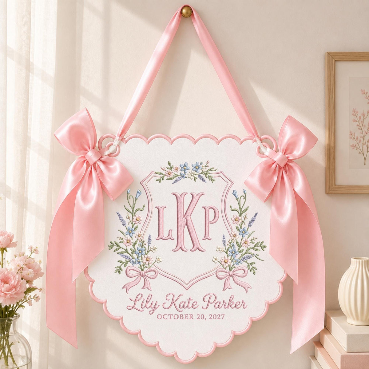 May include: A white scalloped wall hanging featuring pink satin bows on each side and a pink ribbon for suspension. The center displays an embroidered crest with the initials LKP, floral accents, and the text Lily Kate Parker OCTOBER 20, 2027.