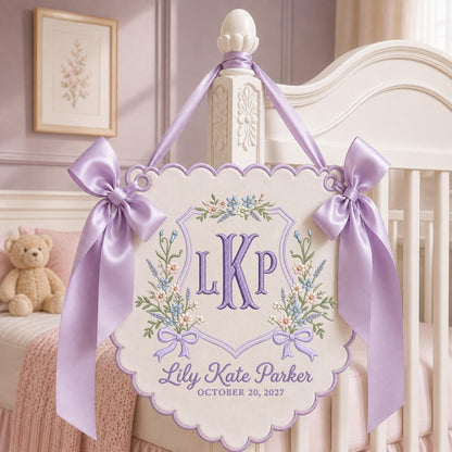 May include: A cream-colored fabric wall hanging with a scalloped edge, featuring purple embroidered monogram letters LKP, floral accents, and the text Lily Kate Parker OCTOBER 20, 2027. It is suspended by a lavender satin ribbon with two large bows.